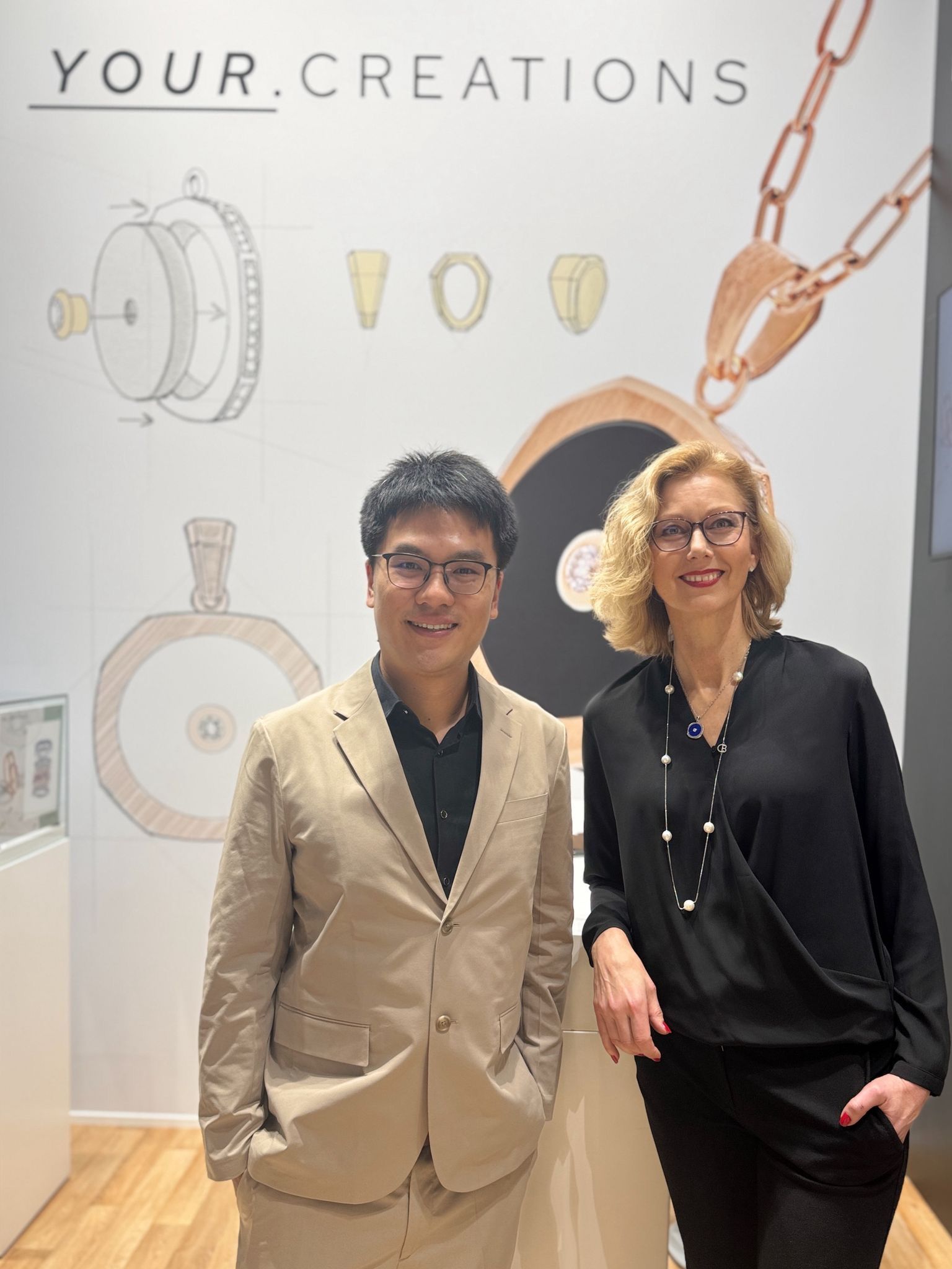 Zwei Personen posieren vor einer Schmuckauslage. Im Hintergrund sind Schmuckdesignskizzen und eine Anhängerabbildung zu sehen.