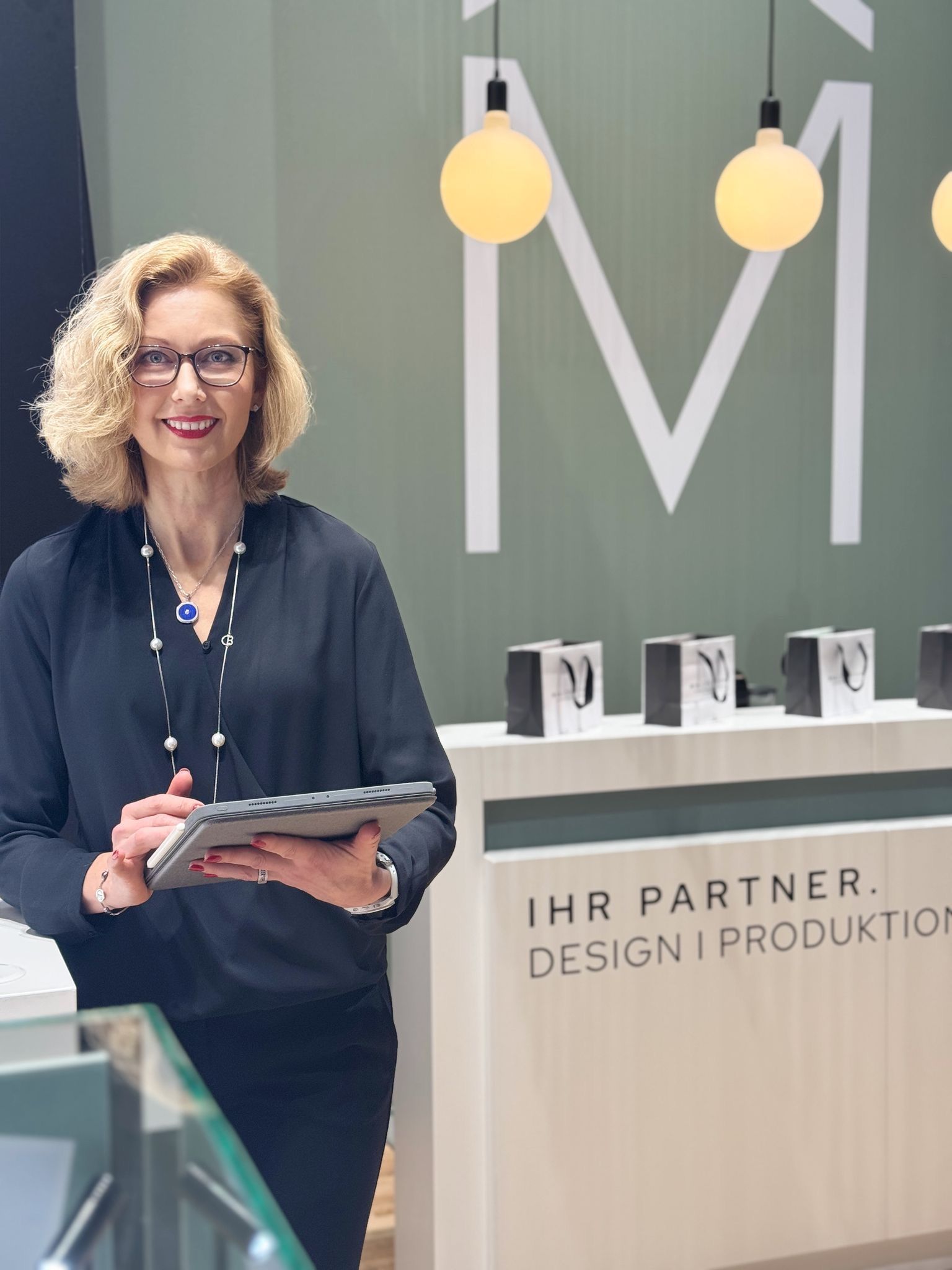 Frau hält ein Tablet in der Hand und lächelt vor einem Display mit Schmuck und Firmenlogo.