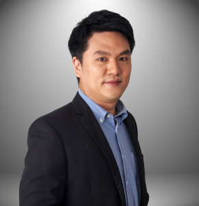 Ein Mann in Anzug und blauem Hemd steht vor einem grauen Hintergrund.