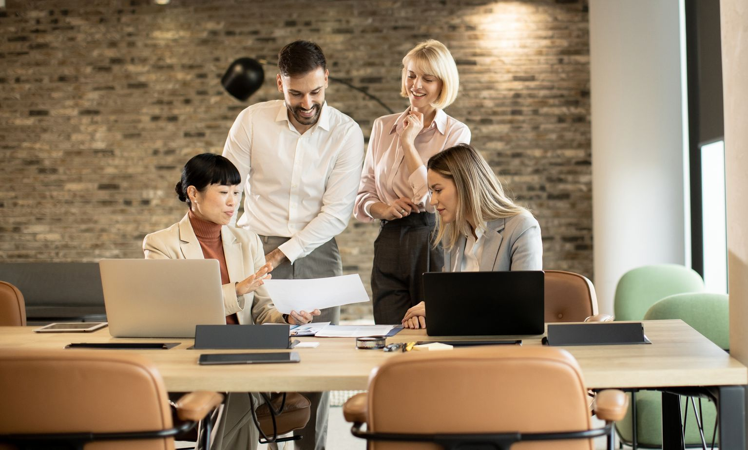 Four colleagues collaborating around a table in an office, reviewing documents and using laptops.