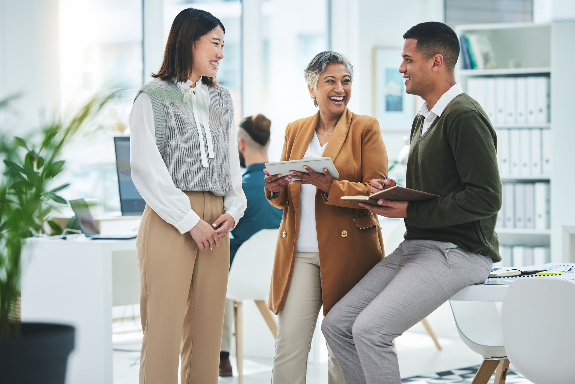 Three colleagues smile and chat while standing in a bright, modern office with a desk and shelving in the background.