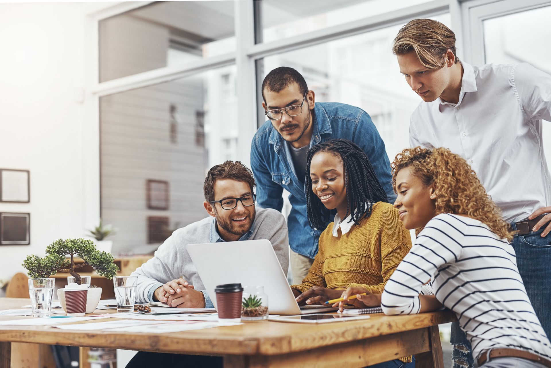 A diverse team collaborates around a laptop on a wooden table in a bright, modern office space.