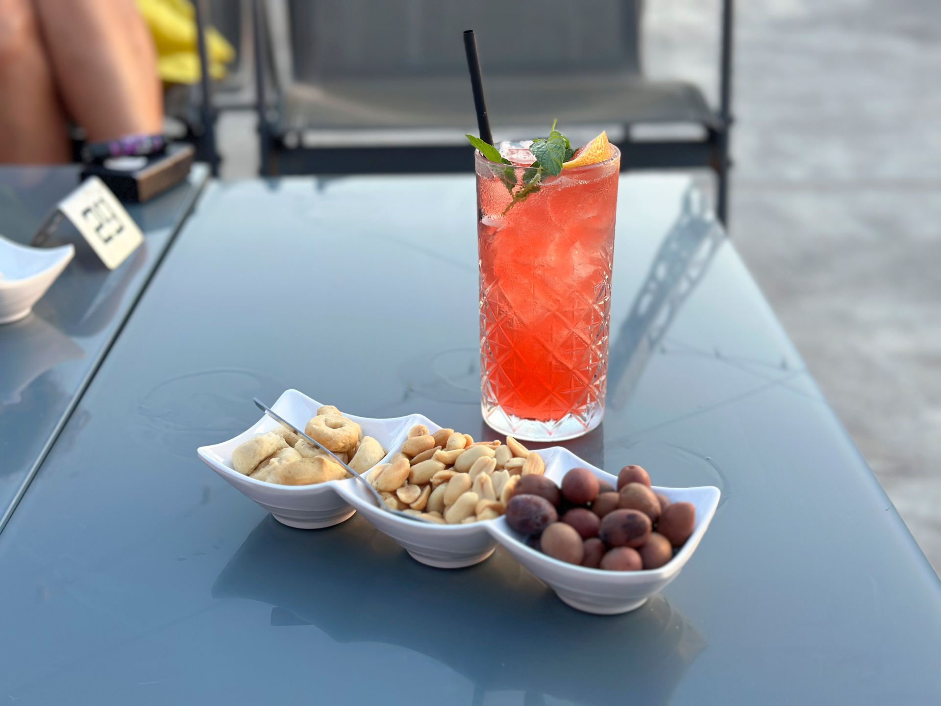 Un bicchiere di liquido rosso accanto a una ciotola di arachidi e olive su un tavolo.