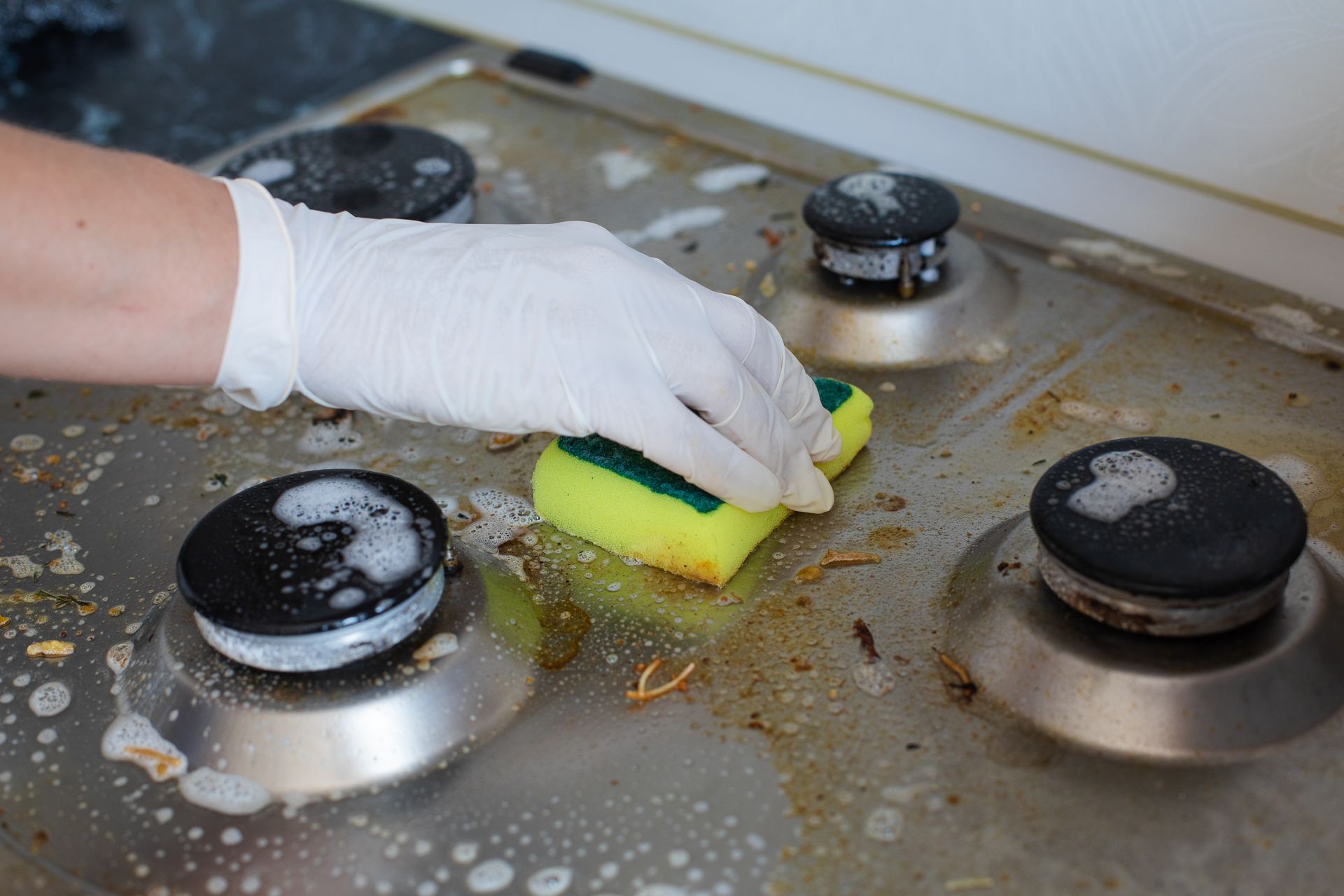 Gloved hand cleaning a dirty stovetop with a sponge.