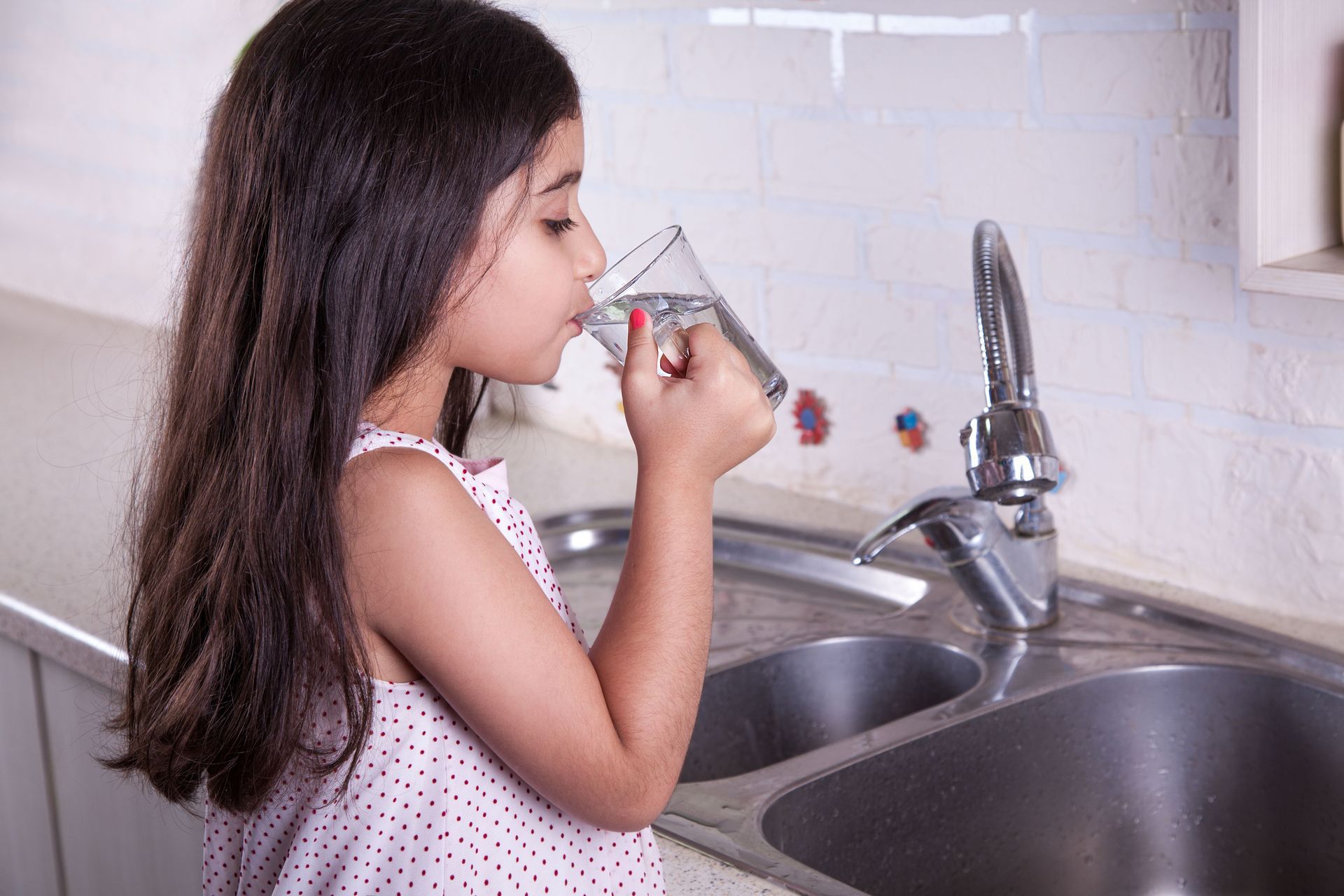 A little girl is drinking water from a glass in a kitchen.