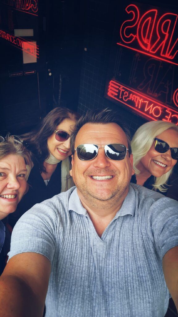 Group selfie: four people smiling, wearing sunglasses. Red neon sign in the background.