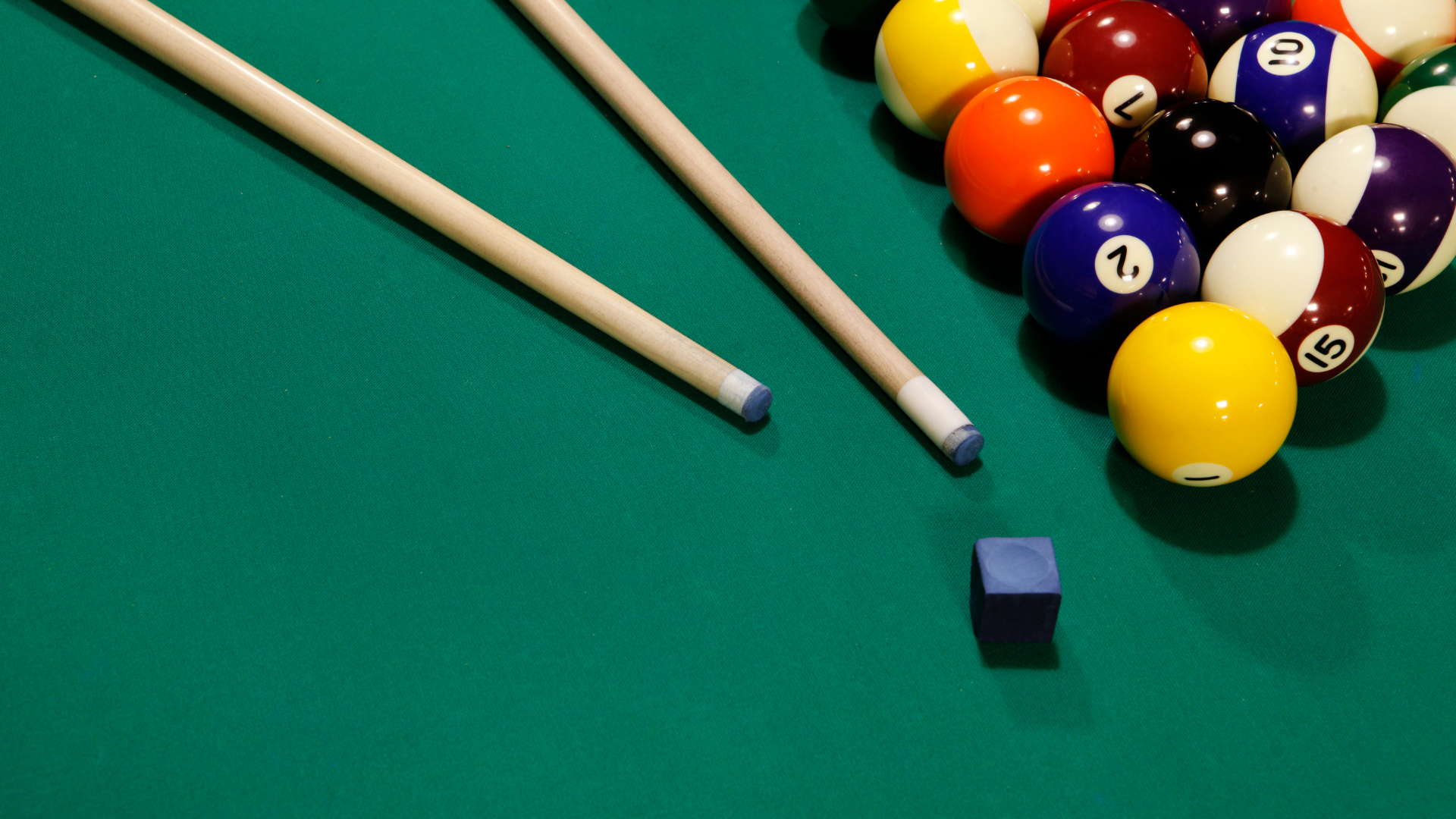Pool table with racked billiard balls, two cues, and a blue chalk cube.