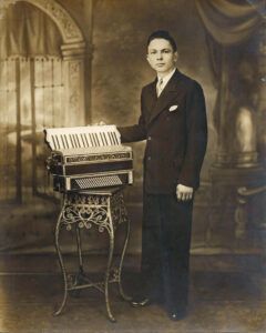 Man in suit poses with an accordion on a decorative stand, in a studio with a painted backdrop.