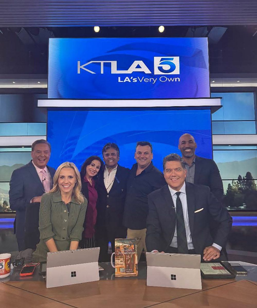 KTLA 5 news team at a desk with a screen in the background. They are smiling in a studio with city backdrop.