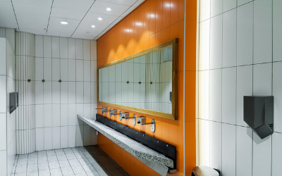 Modern public restroom with white tiled walls, an orange accent wall, a long mirror, and a gray stone trough sink.