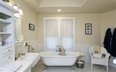 A bright bathroom featuring a white clawfoot bathtub centered under two windows, a vanity, and a white wicker chair.