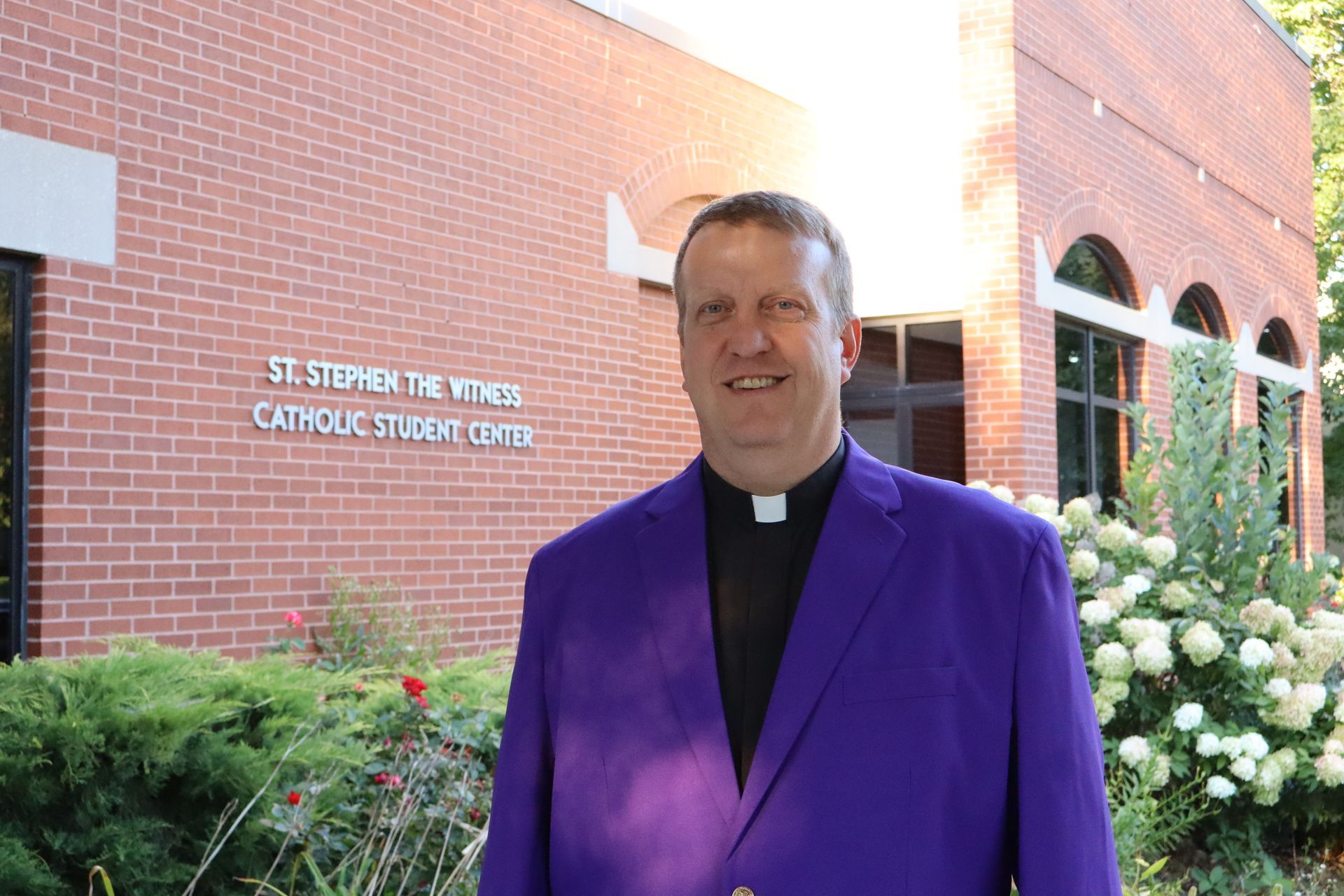 A man in a black suit and tie is smiling in front of a bush of white flowers.