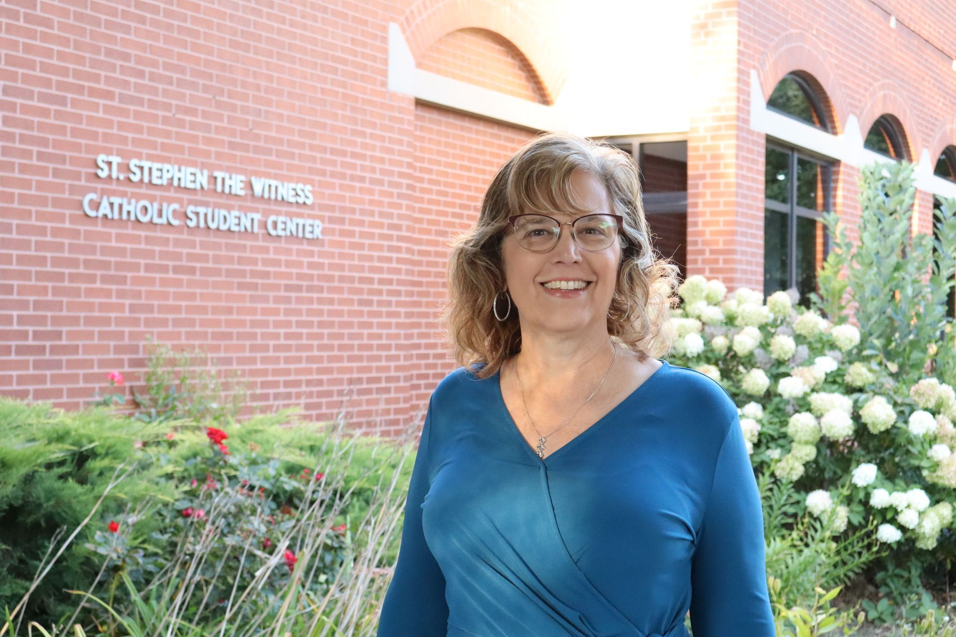A woman wearing glasses and a necklace is smiling in front of a painting.