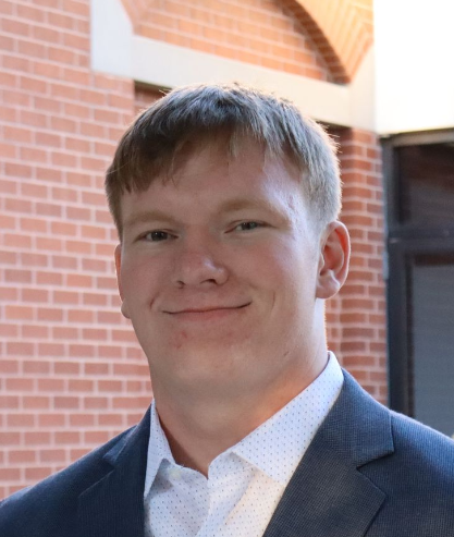 A man in a red shirt is smiling in front of a grey curtain