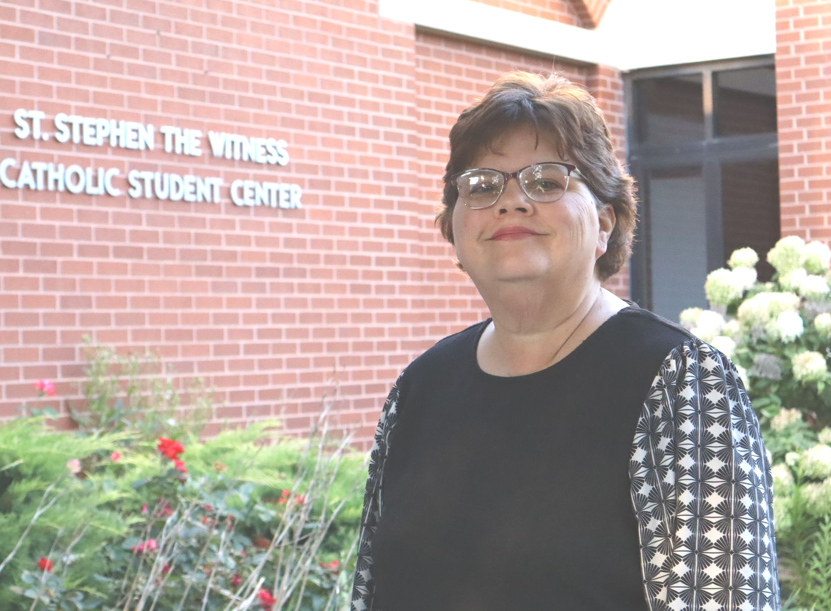 A woman wearing glasses and a striped shirt is smiling in front of a painting.