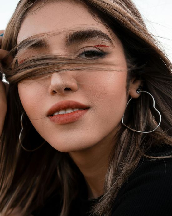 Woman with heart-shaped earrings, holding hair in front of her face. Orange lipstick, wind-blown hair.