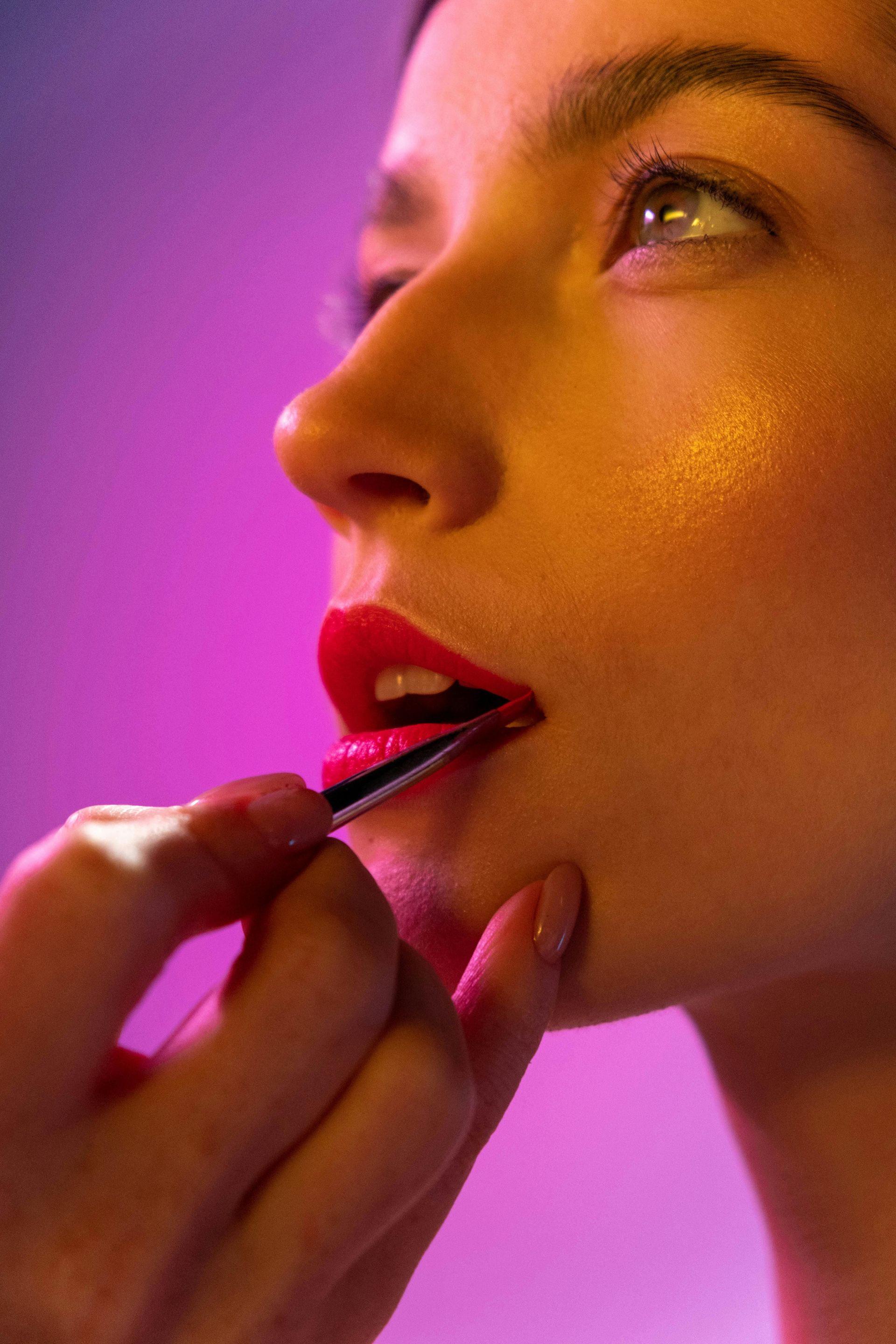 Person's face with red lipstick being applied by hand, against a purple backdrop.