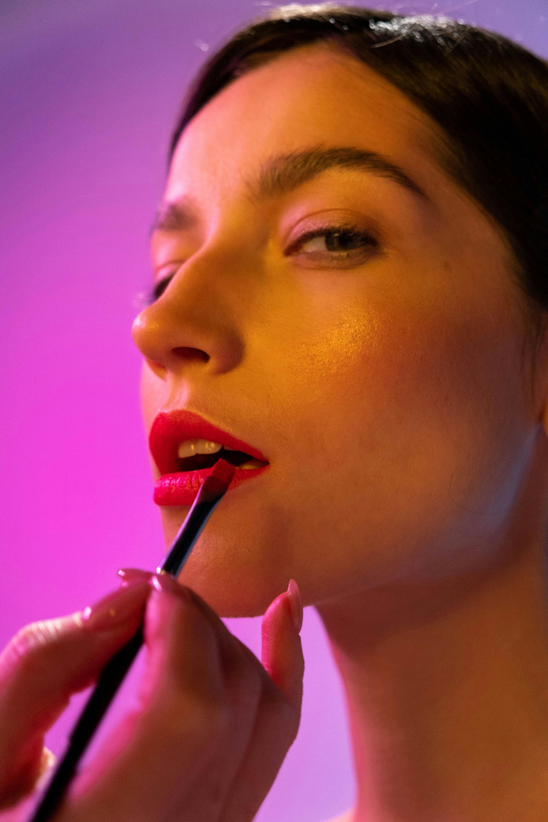 Person applying red lipstick with a brush; bright pink and yellow lighting.