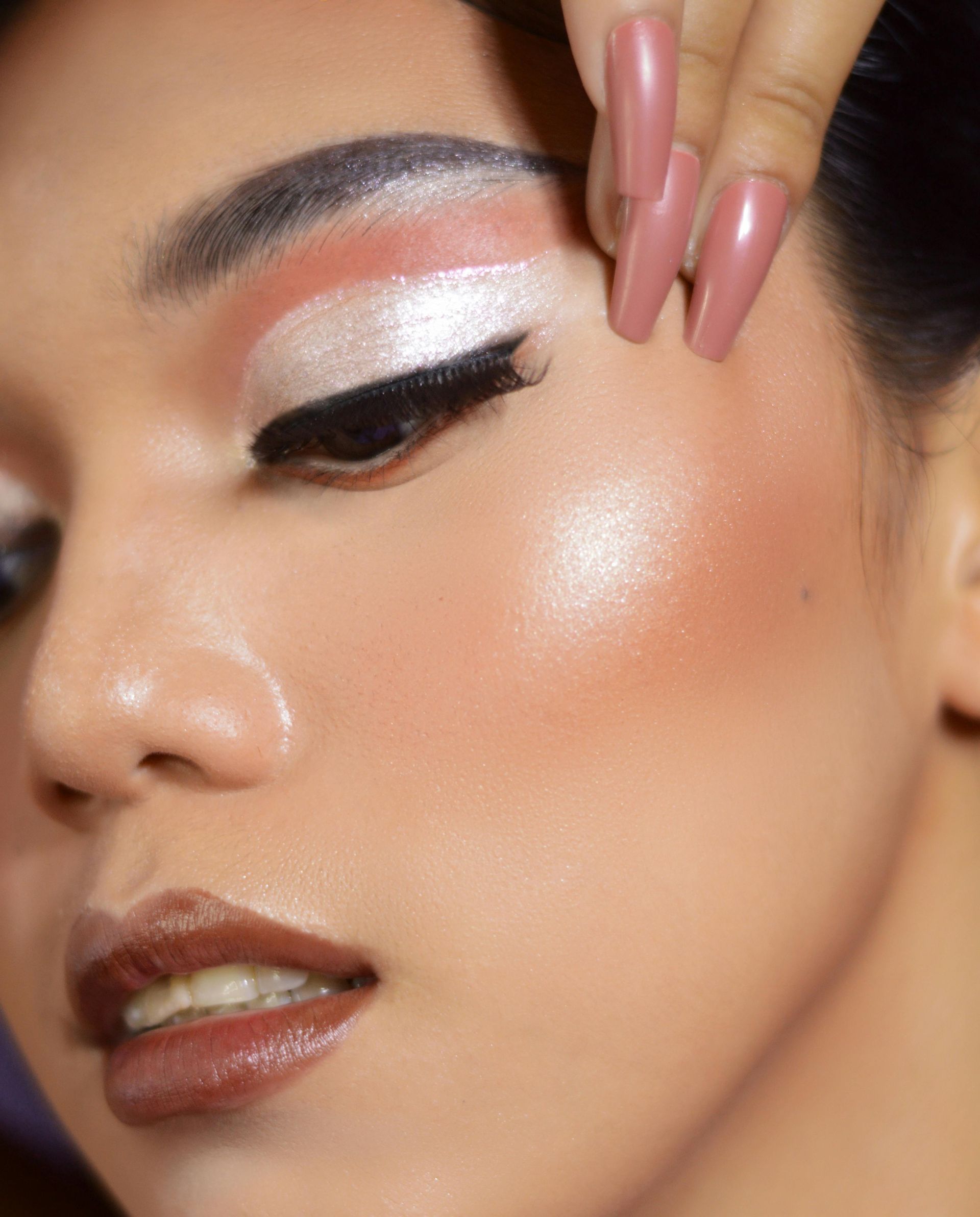 Woman with bold makeup: peach eyeshadow, black winged liner, glossy lips, and long pink nails.