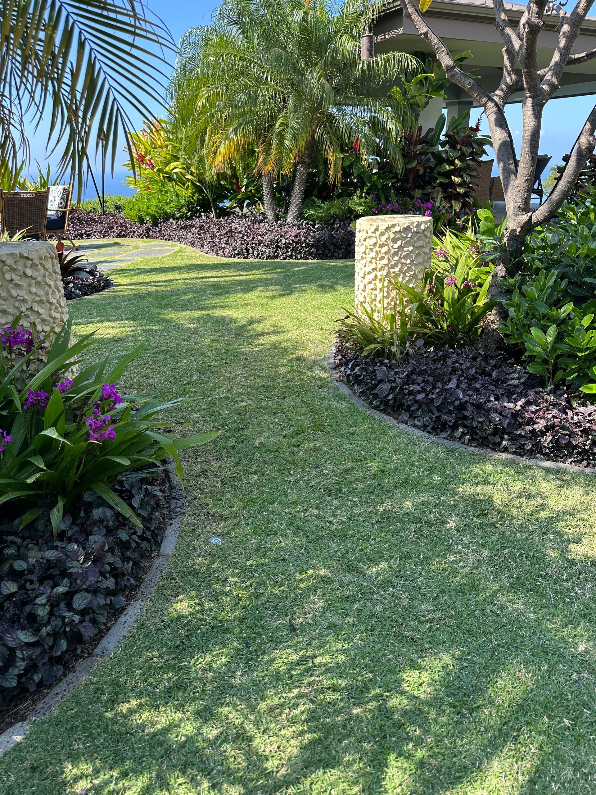 A sunny, manicured garden path with purple plants, green lawn, and stone pillars, leading toward a shaded pavilion.