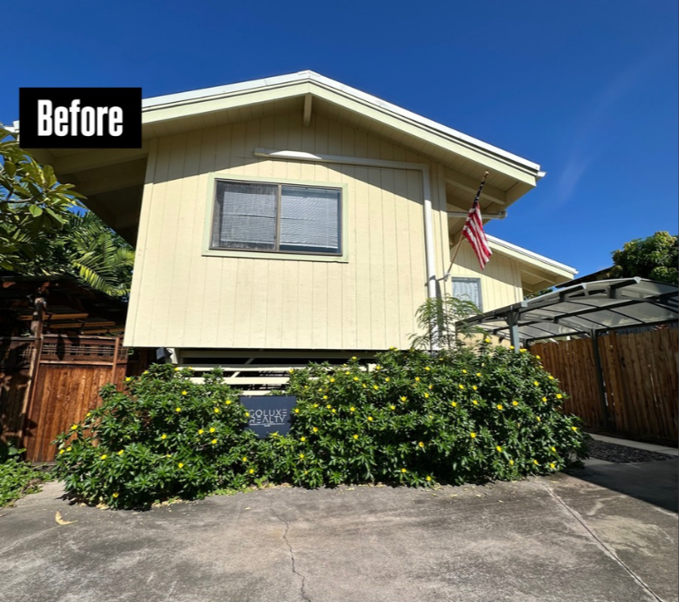 A light yellow house with a gabled roof, an American flag, and a large green bush in the front yard.