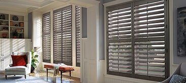 Gray wooden plantation shutters installed on large windows in a sunlit living room with a chair and bench.