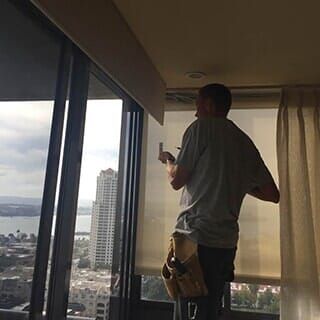 A worker in a gray shirt and tool belt installs a roller shade in front of a window overlooking a city skyline.