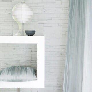 A white shelf against a textured stone wall holds a round lamp and a bowl, with a soft grey curtain to the right.
