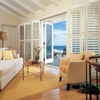 A bright living room with a white sofa, a tan armchair, and folding white shutters opening to an ocean view.