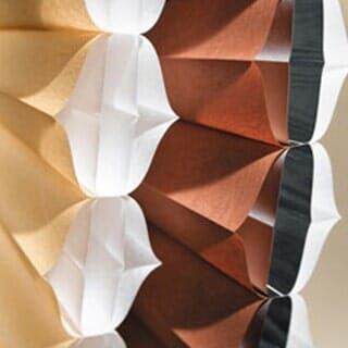 A close-up of accordion-folded paper in shades of white, tan, and rust brown, creating a geometric, textured pattern.