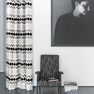 A patterned curtain hangs next to a dark gray chair with a stack of books, under a large portrait on a white wall.