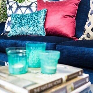 A teal candle holder set rests on stacked books in front of a blue velvet sofa with patterned and pink accent pillows.