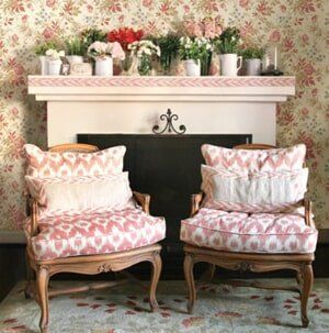 Two floral-patterned armchairs sit before a fireplace mantle decorated with multiple vases of flowers and greenery.