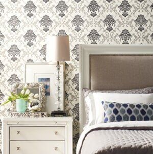 A bedroom featuring damask wallpaper, a white nightstand with a lamp and tulips, and a bed with neutral and blue pillows.