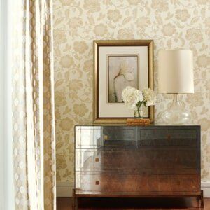 A dark wood dresser topped with a lamp and a framed floral print sits against a wall with cream-colored floral wallpaper.