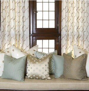A bench seat with various patterned and solid-colored decorative pillows in front of a window with white patterned drapes.