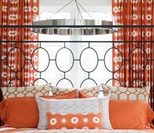 A bed with a dark metal oval-patterned headboard, orange patterned curtains, and decorative throw pillows in orange and tan.