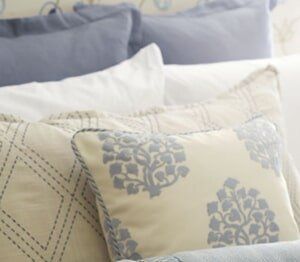 Stacked pillows in shades of white, cream, and soft blue, featuring floral and geometric patterns on a bed.