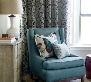 A teal armchair with butterfly and patterned throw pillows sits beside a side table with a lamp and patterned curtains.