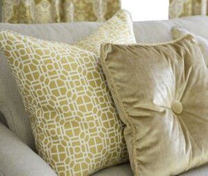 Two throw pillows on a light-colored sofa: one with a gold geometric pattern and one solid gold with a center button.