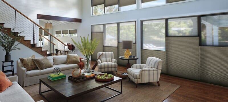 A bright, modern living room with a large sofa, two armchairs, a coffee table, a staircase, and large window shades.