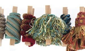 Six wooden clothespins hanging on a string, each holding a different decorative fabric trim or tassel in various colors.