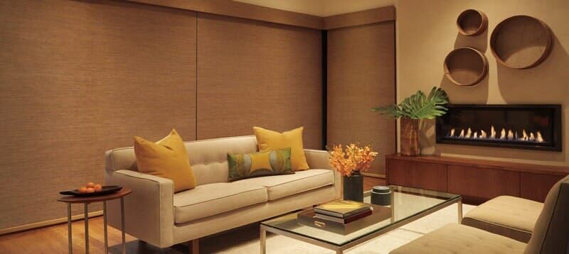 A modern living room with a beige sofa, yellow pillows, a glass coffee table, and a linear fireplace on a brown wall.