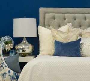 A bed with a tufted gray headboard, white bedding, and decorative pillows against a dark blue wall with a bedside lamp.