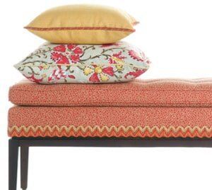 A coral-patterned upholstered bench with a yellow pillow and a floral pillow stacked on top against a white background.