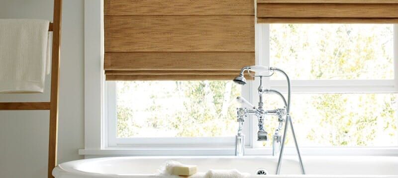 A bathtub with a vintage-style chrome faucet sits beneath a window covered by light brown fabric roman shades.