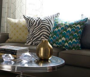 A silver coffee table with a gold vase and books sits before a gray couch featuring yellow, zebra-print, and chevron pillows.