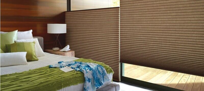 Bedroom with wood walls, green pillows on a bed, and pleated brown shades covering a large window.