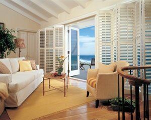 A bright living room with a white sofa, armchair, yellow rug, and floor-to-ceiling shutters opening to an ocean view.