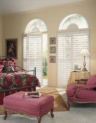 A bedroom features a bed with floral bedding, a pink tufted ottoman, and a matching chair by arched windows with shutters.